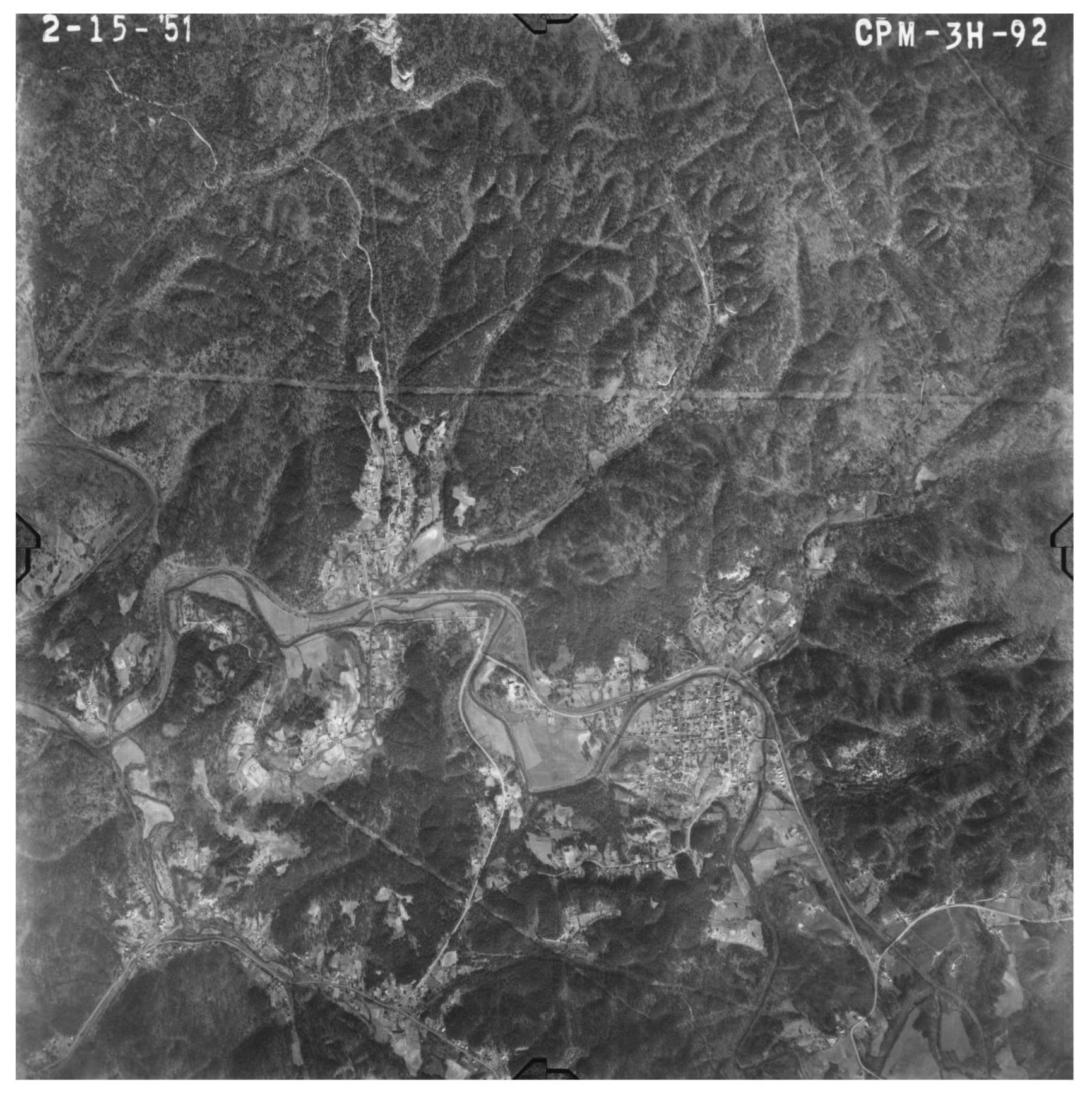 A vertical aerial survey photograph of wooded ridges and creek bottom, the creek looping through open fields, with two clusters of streets and houses down in the valley.